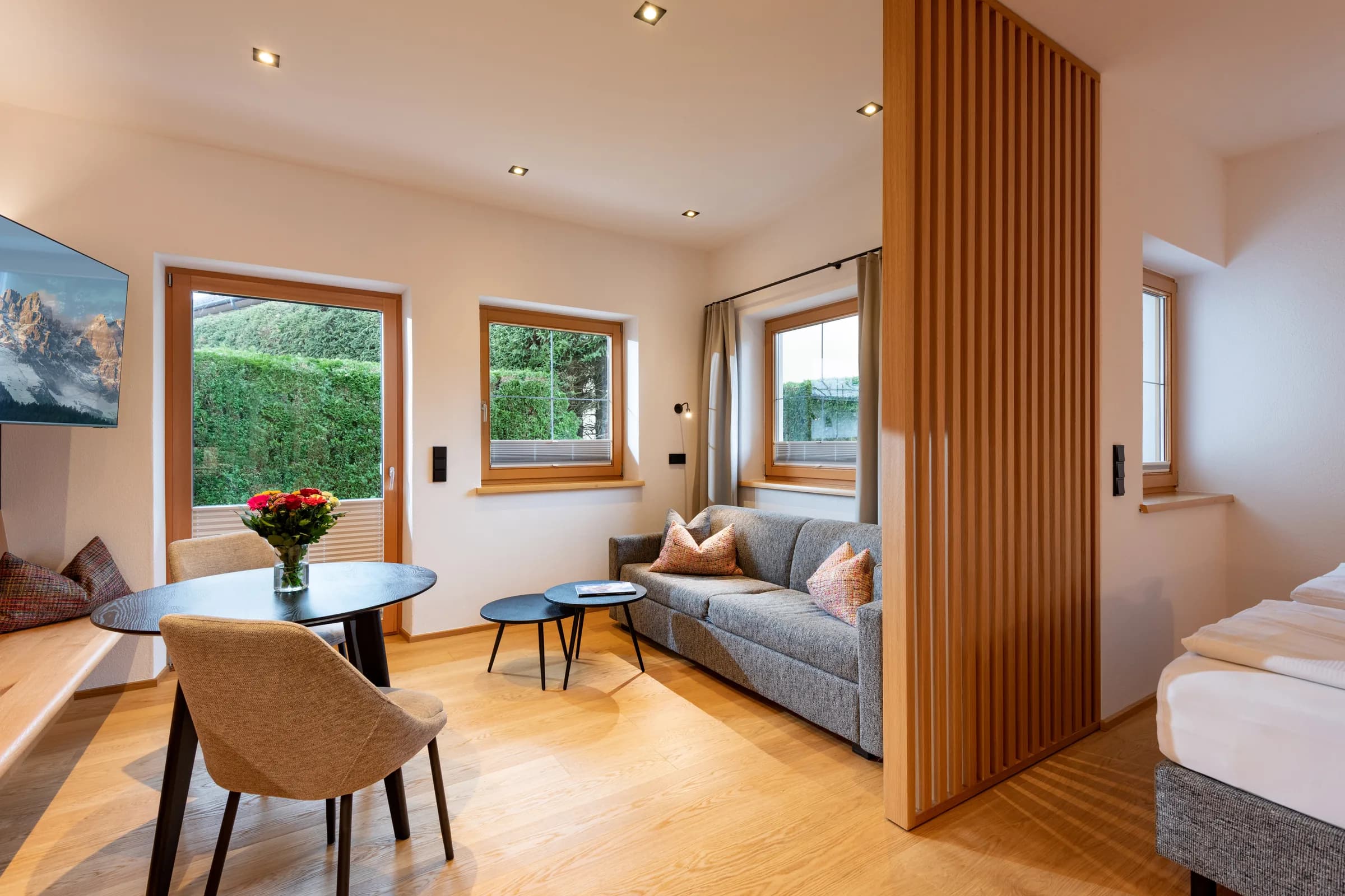 Moderner Wohn- und Schlafbereich mit grauer Couch, Holzlamellen-Raumteiler und Gartenblick im Studio mit Garten des Appartement Erler.