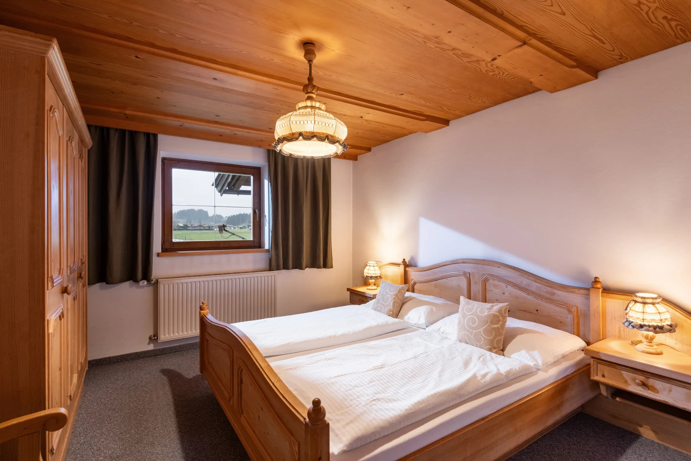 Schlafzimmer mit reich geschnitztem Pinienholz-Doppelbett, Kleiderschrank aus Pinienholz und Fensterblick auf Wiesen und Berge, Appartement Wilder Kaiser.
