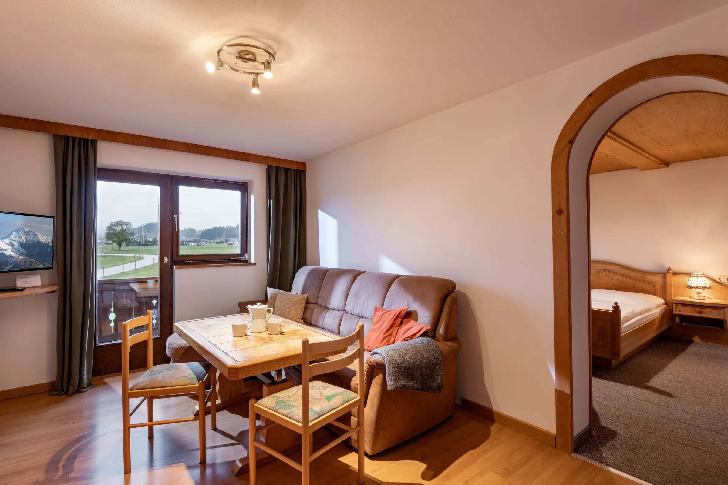 Wohn- und Essbereich mit Couch, Esstisch und Fensterfront mit Blick auf Wiesen und Berge, Bogendurchgang zum Schlafzimmer, Appartement Wilder Kaiser am Bauernhof Oberlacken.