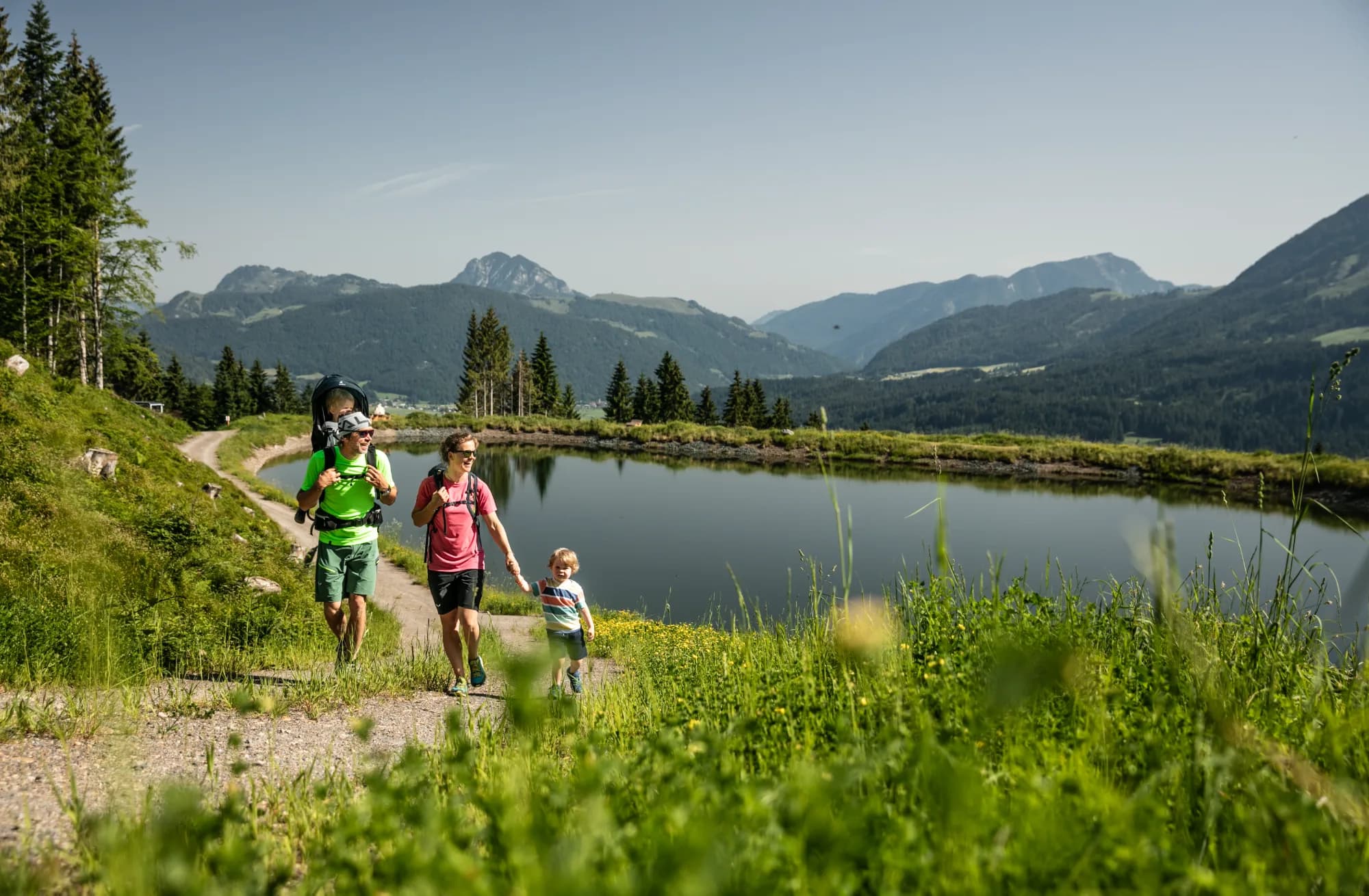 Familie auf einer Sommerwanderung an einem kleinen Bergsee in den Kitzbüheler Alpen, im Hintergrund das weite Tal.