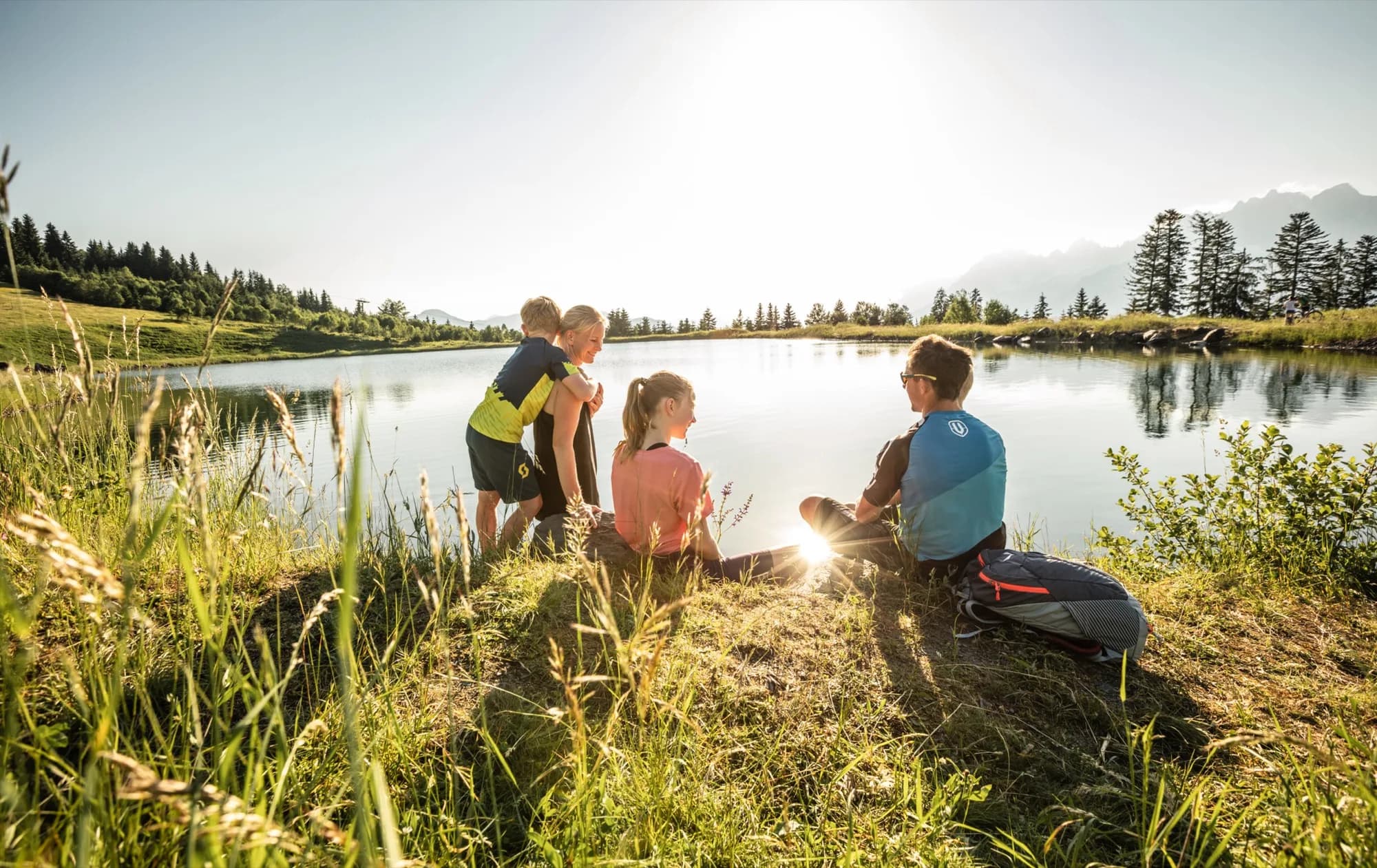 Familie sitzt am Ufer eines Bergsees in den Kitzbüheler Alpen im Abendlicht, im Hintergrund Tannen und die Silhouette der Berge.