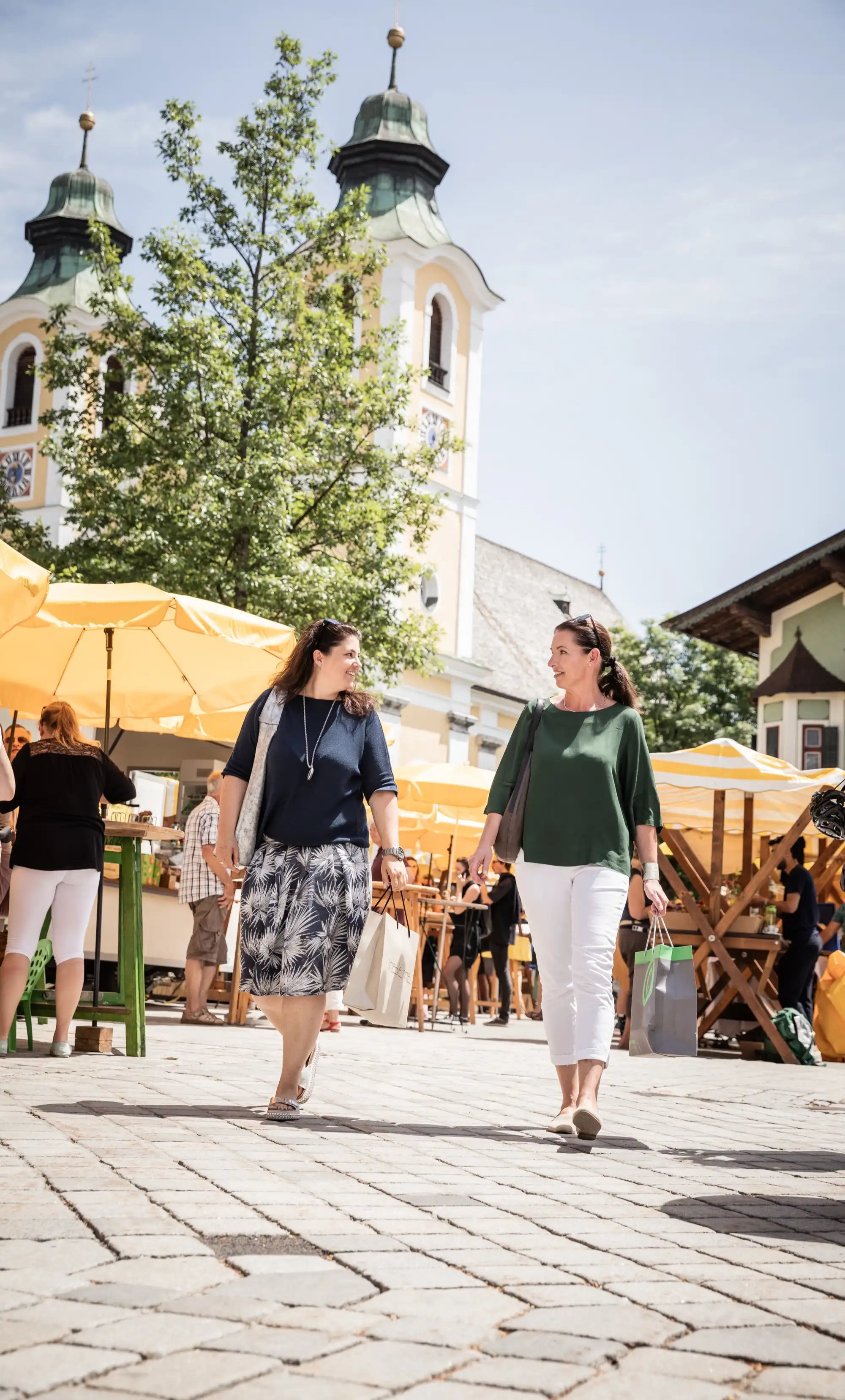 Zwei Frauen schlendern über den Hauptplatz von St. Johann in Tirol; im Hintergrund die Dekanatspfarrkirche und Marktstände mit gelben Sonnenschirmen.