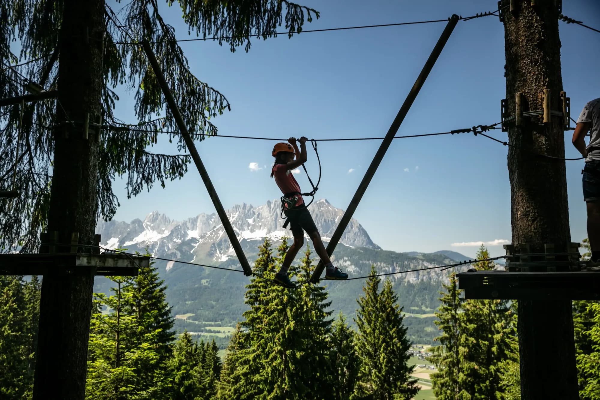 Person auf einem Hochseil-Parcours zwischen den Bäumen im Kletterwald Hornpark, im Hintergrund das Wilder-Kaiser-Massiv.