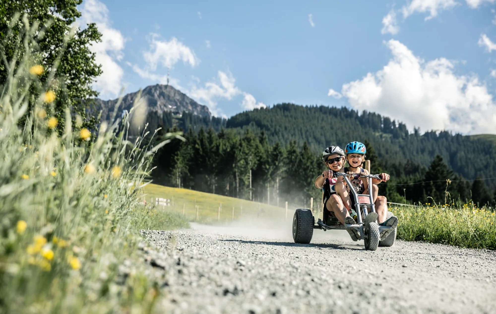 Zwei Kinder rollen mit einem Mountaincart eine Schotterstraße ins Tal, im Hintergrund das Kitzbüheler Horn mit dem Sendeturm.