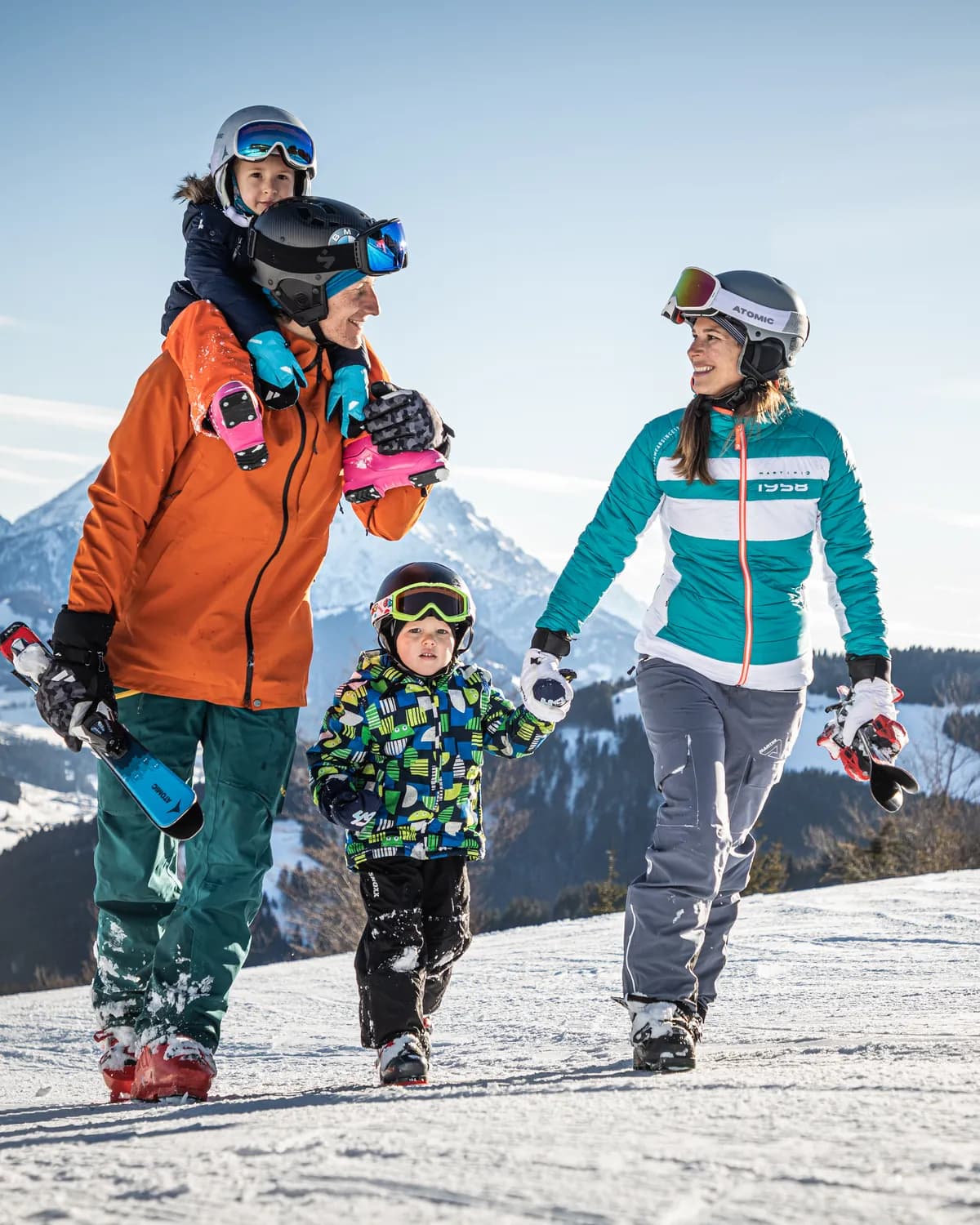 Familie mit zwei kleinen Kindern in Skiausrüstung auf einer Piste, dahinter verschneite Berge.