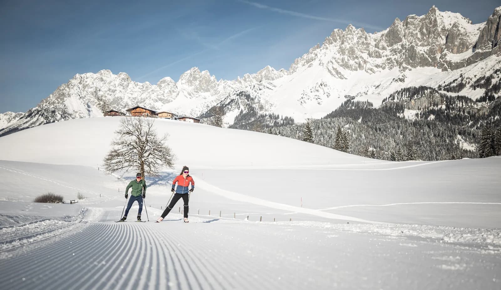Zwei Langläufer auf einer frisch gespurten Loipe bei St. Johann in Tirol, dahinter Bauernhöfe am Hang und der verschneite Wilde Kaiser.
