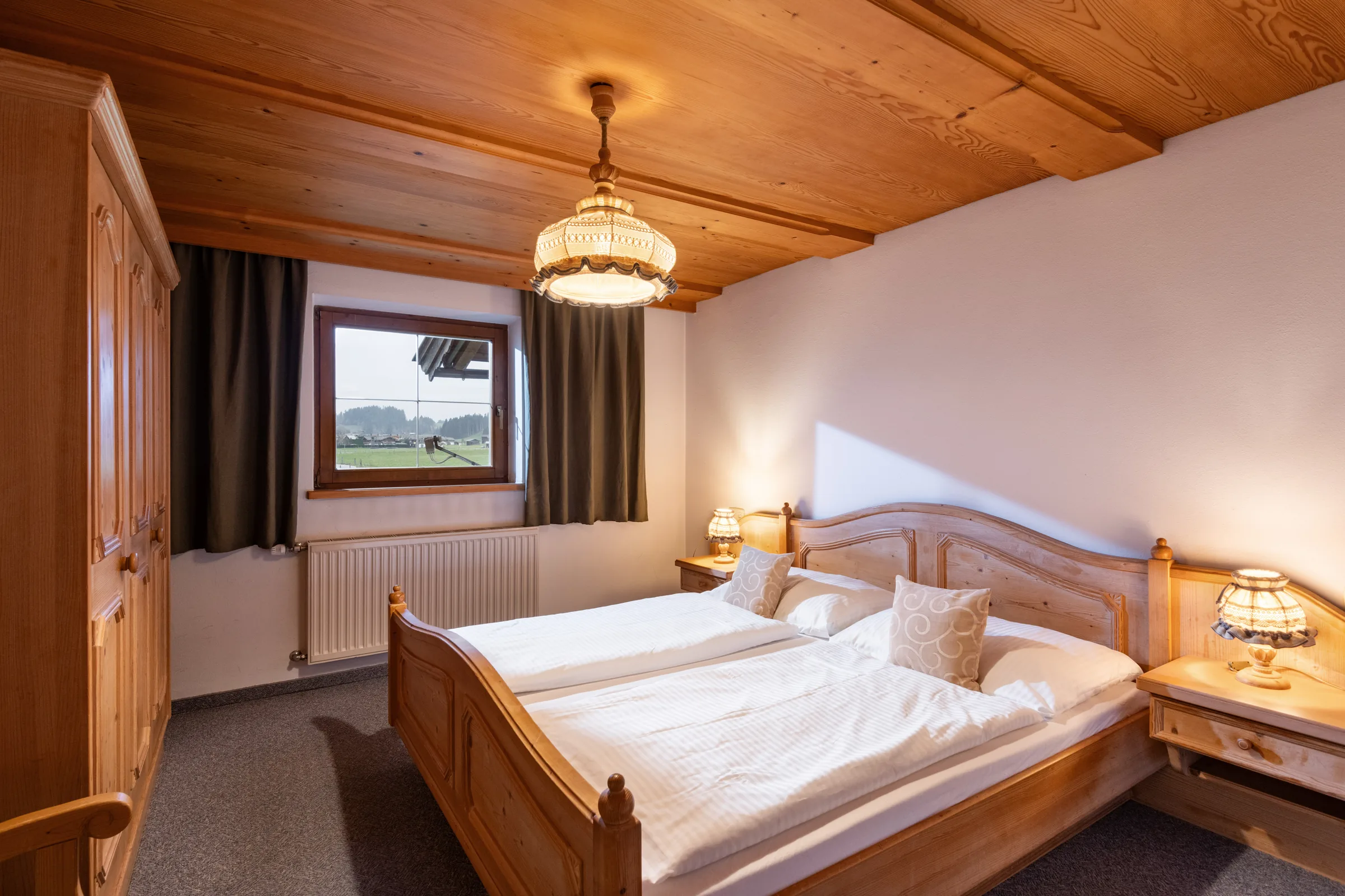Schlafzimmer mit reich geschnitztem Pinienholz-Doppelbett und Fensterblick auf Wiesen und Berge, Appartement Wilder Kaiser am Bauernhof Oberlacken.